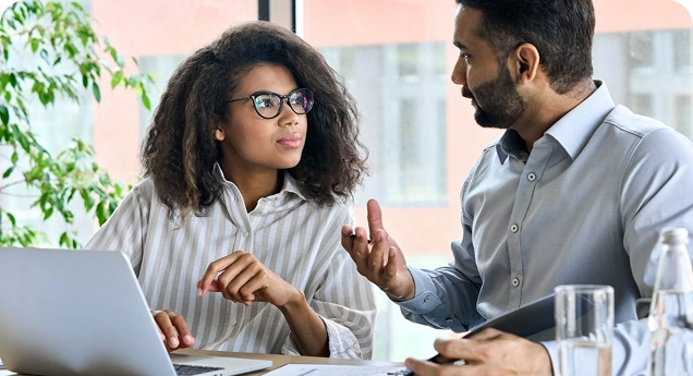 Duas pessoas sentadas à mesa em um ambiente de trabalho, conversando. À esquerda, uma mulher de pele escura, com cabelo cacheado volumoso e óculos, olha atentamente para a pessoa ao lado enquanto usa um notebook aberto à sua frente. À direita, um homem de pele clara, com barba curta e camisa social, gesticula com uma das mãos enquanto fala. Sobre a mesa há papéis e um copo de água. O ambiente é iluminado por luz natural, com uma planta e janelas ao fundo, sugerindo um escritório ou sala de reunião.
