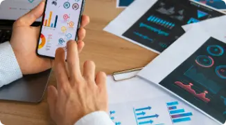 Mãos de uma pessoa mexendo em um celular com gráficos variados e papéis em volta cheios de gráficos.